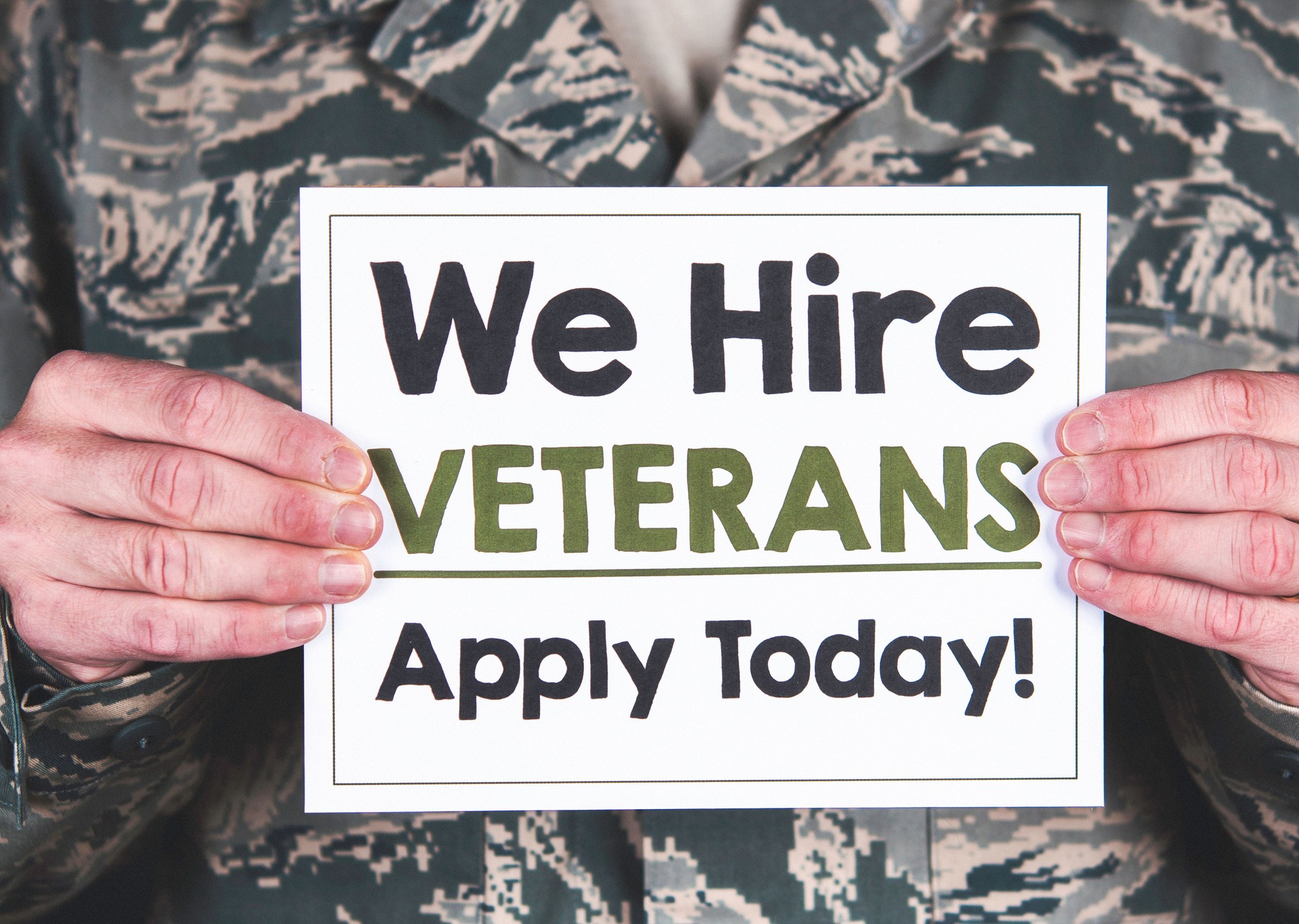 American soldier in uniform holding a sign that reads We Hire Veterans